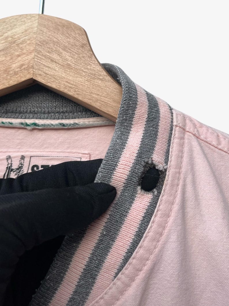 Puede incluir: Primer plano de un cuello de chaqueta rosa con ribete a rayas grises y rosas. La chaqueta est&aacute; abotonada con un bot&oacute;n negro. El cuello est&aacute; en una percha de madera.