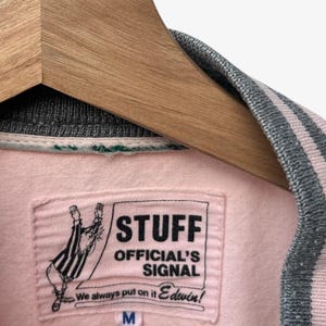 Puede incluir: Chaqueta universitaria rosa y gris con una etiqueta que dice "STUFF OFFICIAL'S SIGNAL" y "Edwin!". La chaqueta est&aacute; colgada en una percha de madera. El cuello y los pu&ntilde;os tienen rayas grises y rosas.