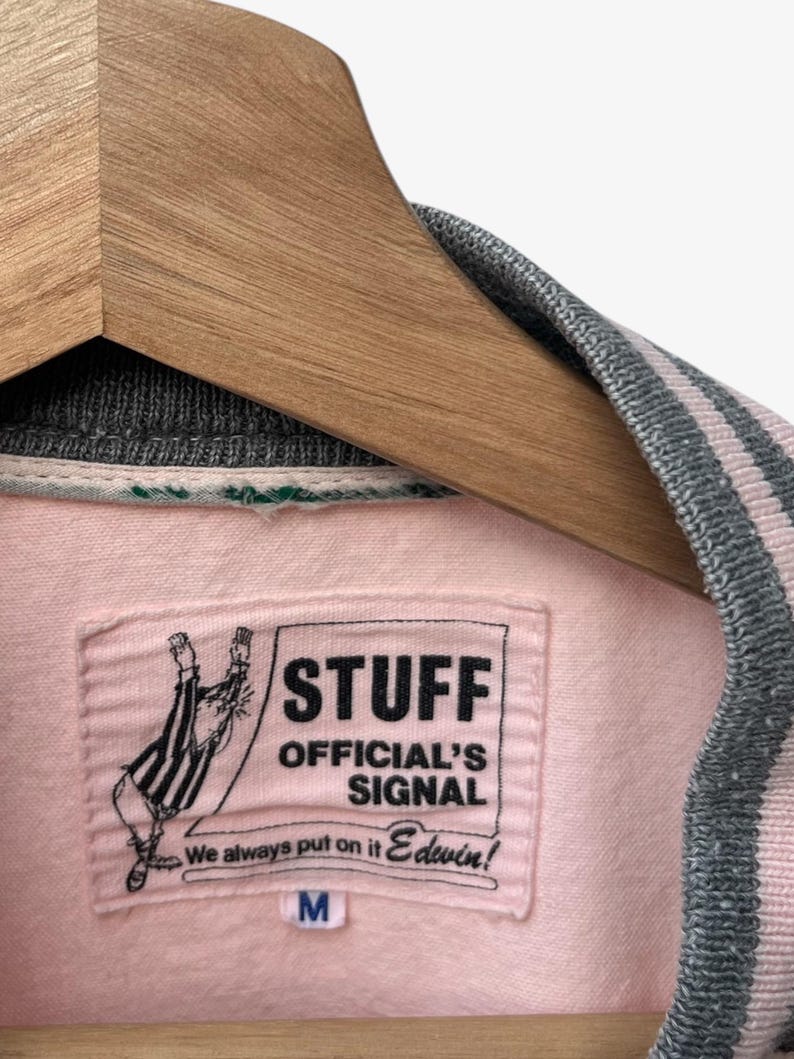 Puede incluir: Chaqueta universitaria rosa y gris con una etiqueta que dice "STUFF OFFICIAL'S SIGNAL" y "Edwin!". La chaqueta est&aacute; colgada en una percha de madera. El cuello y los pu&ntilde;os tienen rayas grises y rosas.