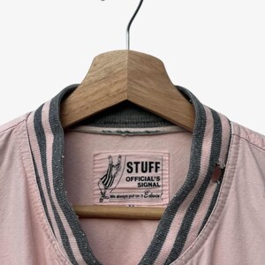 Puede incluir: Chaqueta bomber rosa claro con cuello y pu&ntilde;os a rayas grises y rosas, colgada de una percha de madera. La chaqueta presenta una etiqueta con el texto "STUFF OFFICIAL'S SIGNAL" y un gr&aacute;fico de una mano.
