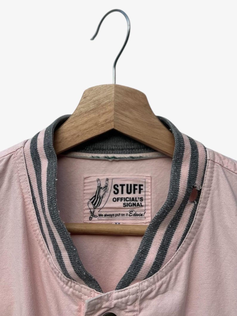 Puede incluir: Chaqueta bomber rosa claro con cuello y pu&ntilde;os a rayas grises y rosas, colgada de una percha de madera. La chaqueta presenta una etiqueta con el texto "STUFF OFFICIAL'S SIGNAL" y un gr&aacute;fico de una mano.