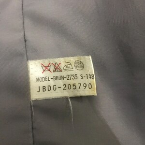 May include: Close-up of a gray fabric with a white tag attached. The tag has text and symbols, including "MODEL-BRON-2735 S-148" and "JBDG-205790". The fabric appears to be part of a garment.