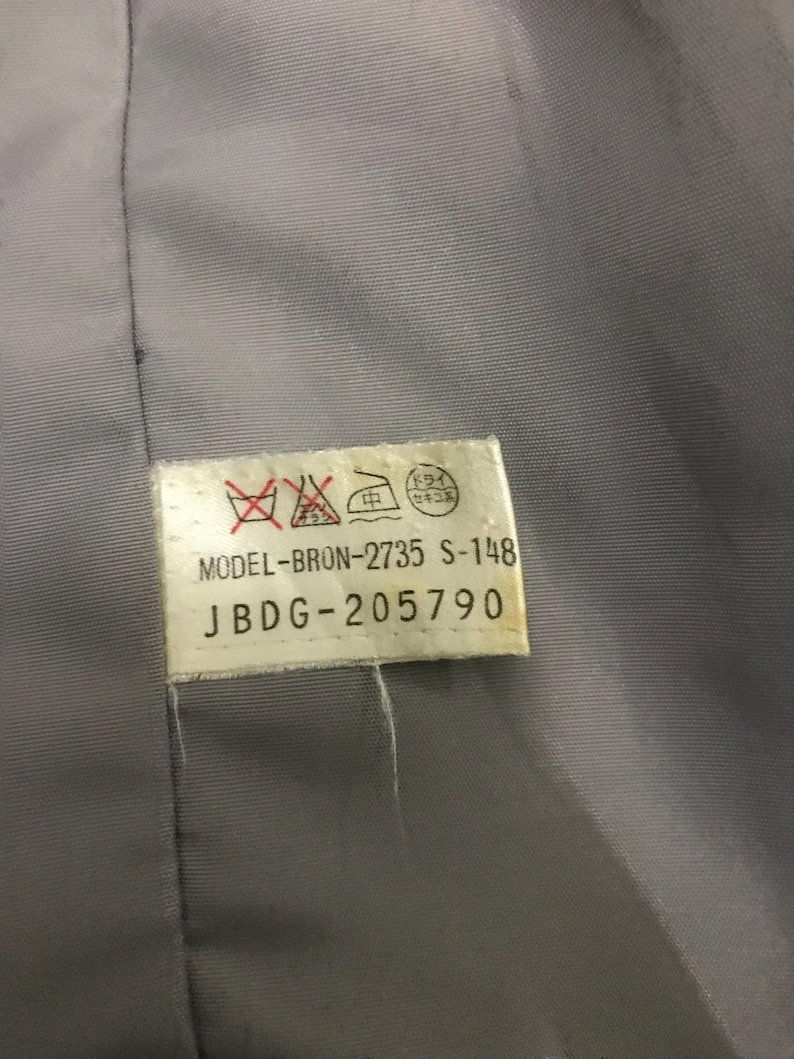 May include: Close-up of a gray fabric with a white tag attached. The tag has text and symbols, including "MODEL-BRON-2735 S-148" and "JBDG-205790". The fabric appears to be part of a garment.