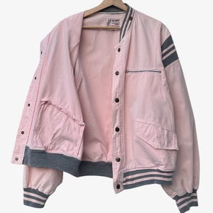 Puede incluir: Chaqueta bomber rosa con detalles grises, incluyendo rayas en las mangas y pu&ntilde;os. La chaqueta tiene un bolsillo con cremallera y cierres de bot&oacute;n. La palabra "STUFF" es visible en la etiqueta.
