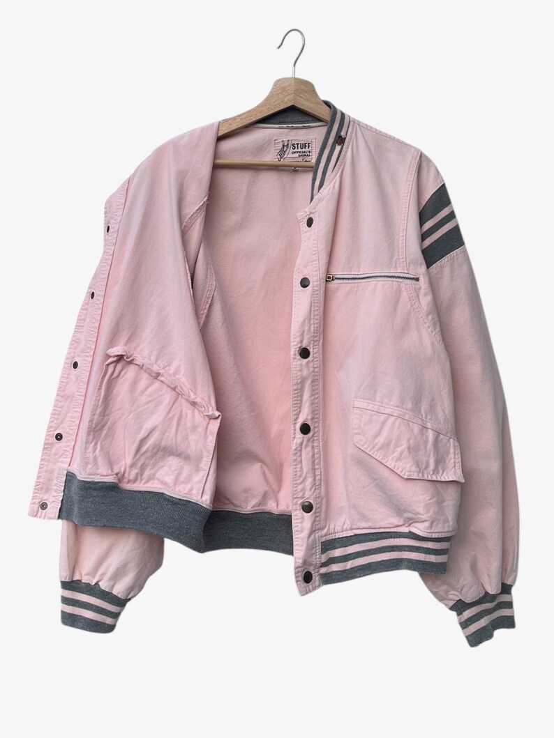 Puede incluir: Chaqueta bomber rosa con detalles grises, incluyendo rayas en las mangas y pu&ntilde;os. La chaqueta tiene un bolsillo con cremallera y cierres de bot&oacute;n. La palabra "STUFF" es visible en la etiqueta.