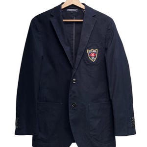 Könnte beinhalten: Marineblauer Blazer mit Zwei-Knopf-Verschluss, aufgesetzten Taschen und einem kleinen gestickten Emblem auf der Brust. Der Blazer hängt an einem Holzkleiderbügel. Das Emblem zeigt den Text "HILFIGER" und "85" in einem Schild.