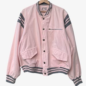 Puede incluir: Chaqueta bomber rosa con rayas grises en las mangas y el cuello. La chaqueta tiene un bolsillo con cremallera en el pecho y dos bolsillos delanteros. Cuelga de una percha de madera.