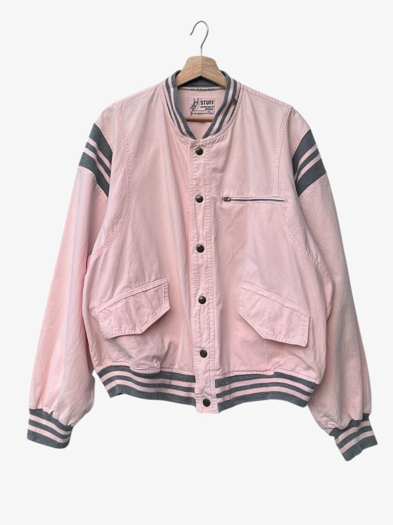 Puede incluir: Chaqueta bomber rosa con rayas grises en las mangas y el cuello. La chaqueta tiene un bolsillo con cremallera en el pecho y dos bolsillos delanteros. Cuelga de una percha de madera.