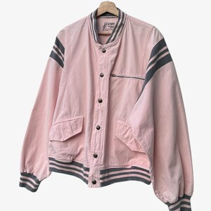 Puede incluir: Chaqueta bomber rosa con rayas grises en las mangas y el dobladillo. La chaqueta tiene un bolsillo con cremallera, cierre de botones y la palabra "STUFF" en la etiqueta. Cuelga de una percha de madera.