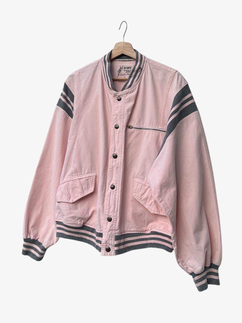 Puede incluir: Chaqueta bomber rosa con rayas grises en las mangas y el dobladillo. La chaqueta tiene un bolsillo con cremallera, cierre de botones y la palabra "STUFF" en la etiqueta. Cuelga de una percha de madera.