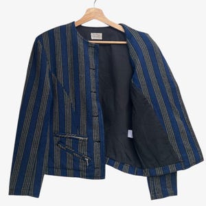 May include: A blue and gray striped jacket with a black interior, hanging on a wooden hanger. The jacket has two zippered pockets and a button closure. The jacket is open, revealing the black lining.