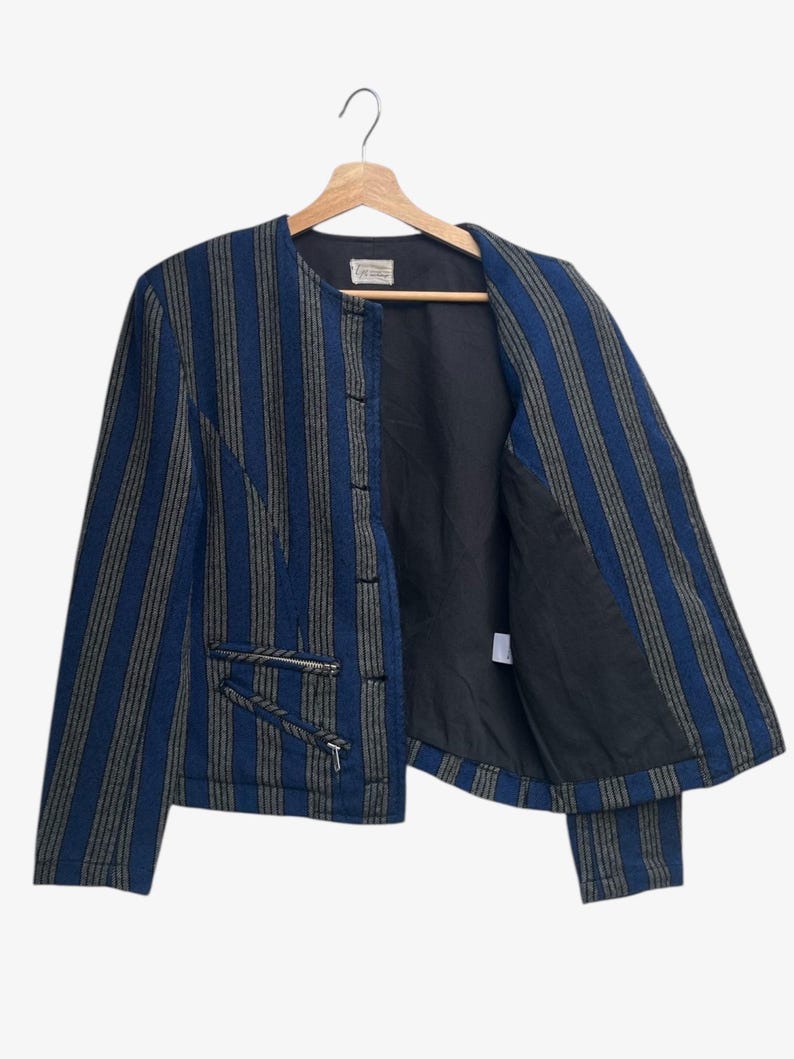 May include: A blue and gray striped jacket with a black interior, hanging on a wooden hanger. The jacket has two zippered pockets and a button closure. The jacket is open, revealing the black lining.