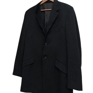 K&ouml;nnte beinhalten: Ein schwarzer Blazer mit fallendem Revers, zwei Vordertaschen und einem Zwei-Knopf-Verschluss. Der Blazer h&auml;ngt an einem Holzkleiderb&uuml;gel vor wei&szlig;em Hintergrund. Das Kleidungsst&uuml;ck hat ein klassisches Design.