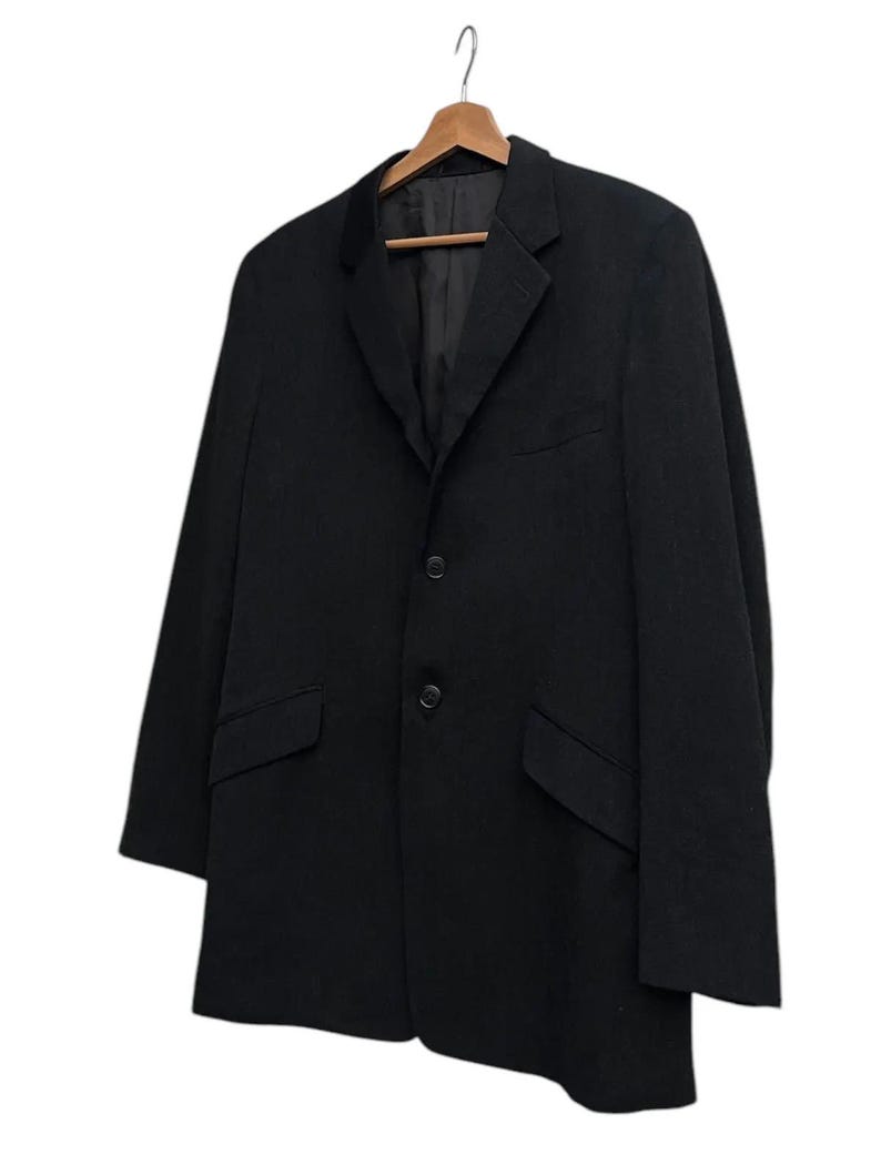 K&ouml;nnte beinhalten: Ein schwarzer Blazer mit fallendem Revers, zwei Vordertaschen und einem Zwei-Knopf-Verschluss. Der Blazer h&auml;ngt an einem Holzkleiderb&uuml;gel vor wei&szlig;em Hintergrund. Das Kleidungsst&uuml;ck hat ein klassisches Design.