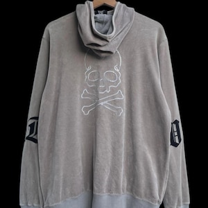 May include: A taupe hoodie with a skull and crossbones design on the back. The skull is outlined in white stitching. The sleeves have a black and white letter design. The hoodie has a gray hood and trim.