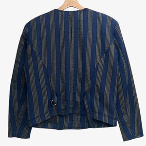 May include: A blue and gray striped jacket with long sleeves, hanging on a wooden hanger. The jacket has a round neckline and a metal ring detail near the waist. The stripes run vertically.