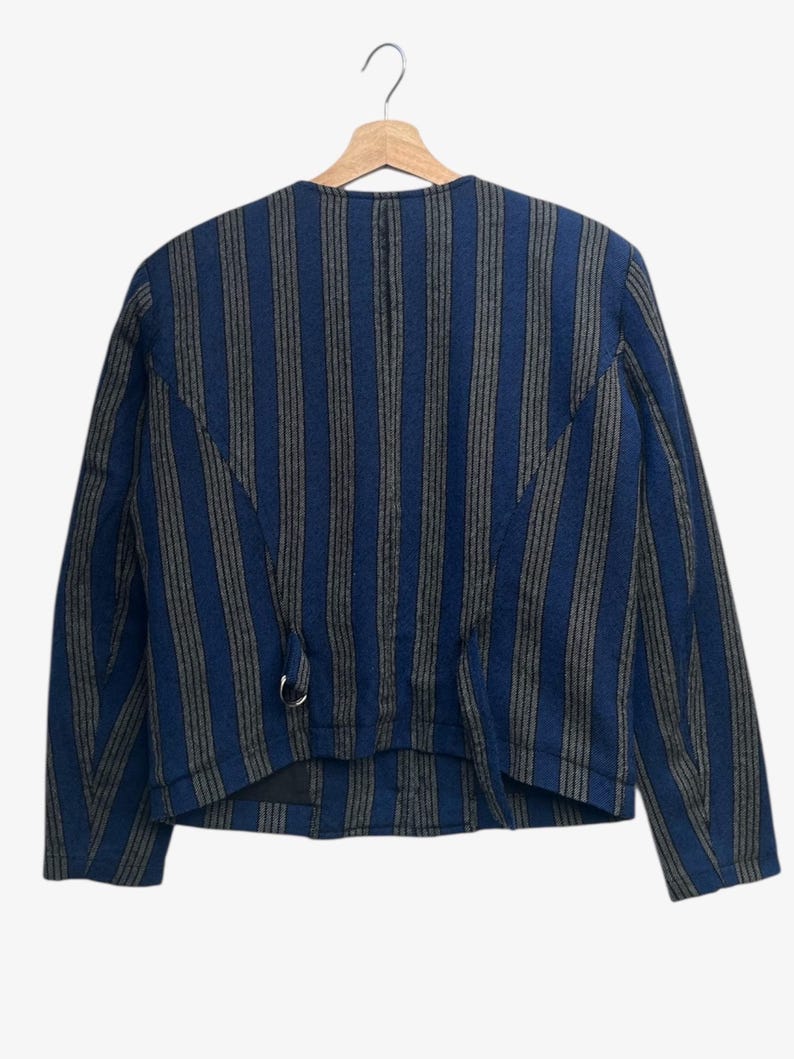 May include: A blue and gray striped jacket with long sleeves, hanging on a wooden hanger. The jacket has a round neckline and a metal ring detail near the waist. The stripes run vertically.