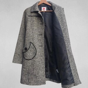 May include: A gray and white tweed coat with a black trim and a black interior lining. The coat has a unique pocket design with a black outline and a large black button. The coat is hanging on a wooden hanger.