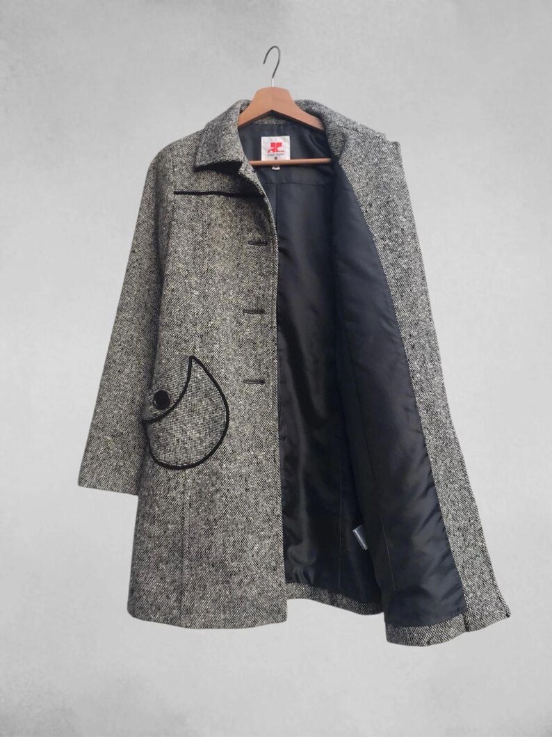 May include: A gray and white tweed coat with a black trim and a black interior lining. The coat has a unique pocket design with a black outline and a large black button. The coat is hanging on a wooden hanger.