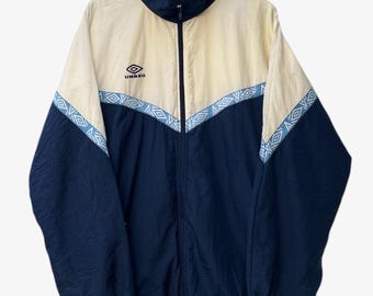 Puede incluir: Chaqueta deportiva Umbro vintage, azul marino y crema. La chaqueta tiene cremallera completa, cuello alto y diseño geométrico azul claro en el pecho y mangas. El logotipo de Umbro es visible.