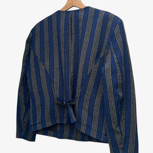 May include: A blue and gray striped jacket hangs on a wooden hanger. The jacket has a tailored fit with a small tie detail at the back. The stripes run vertically, creating a classic and versatile look.