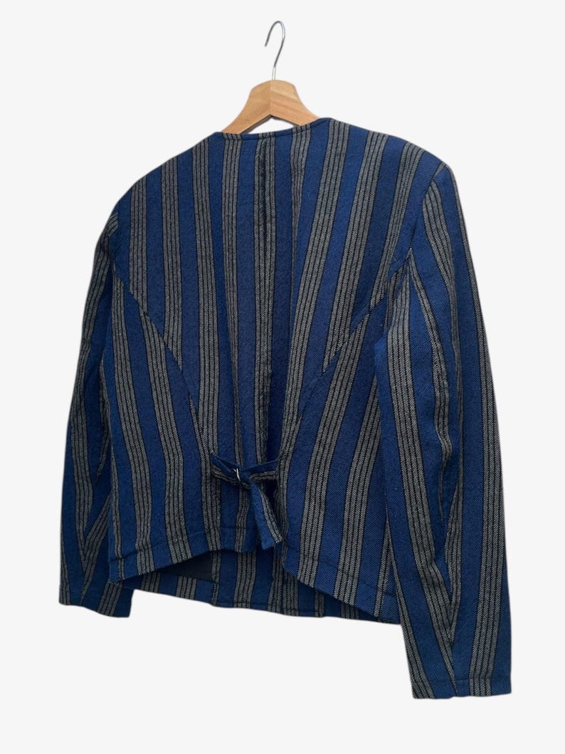 May include: A blue and gray striped jacket hangs on a wooden hanger. The jacket has a tailored fit with a small tie detail at the back. The stripes run vertically, creating a classic and versatile look.