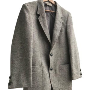 May include: A gray tweed blazer with a two-button closure, two front pockets, and buttons on the sleeves. The blazer is hanging on a wooden hanger against a white background. The fabric has a subtle, textured pattern.