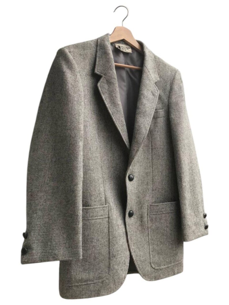 May include: A gray tweed blazer with a two-button closure, two front pockets, and buttons on the sleeves. The blazer is hanging on a wooden hanger against a white background. The fabric has a subtle, textured pattern.