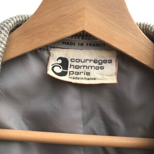 May include: Close-up of a vintage jacket label. The label reads "courr&egrave;ges hommes paris made in france". The jacket is hanging on a wooden hanger. The jacket's interior lining is gray.