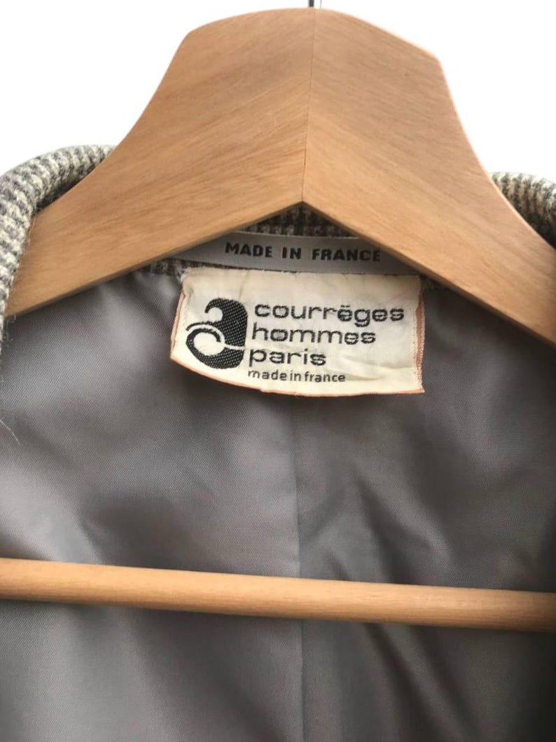 May include: Close-up of a vintage jacket label. The label reads "courr&egrave;ges hommes paris made in france". The jacket is hanging on a wooden hanger. The jacket's interior lining is gray.