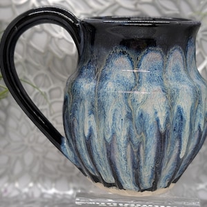 Puede incluir: Una taza de cerámica hecha a mano con asa y borde negros. El cuerpo de la taza presenta un esmalte azul y blanco con efecto goteado. La taza está diseñada para bebidas calientes.