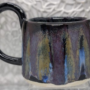 May include: A ceramic mug featuring a glossy black handle and a cylindrical body with a reactive glaze finish. The exterior displays vertical streaks of deep purple, metallic gold, and vibrant blue tones over a dark base, with a plain white base.