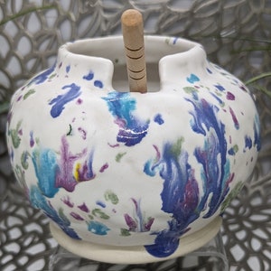 May include: White ceramic yarn bowl with a wooden spindle, featuring a colorful abstract design with blue, purple, green, and yellow accents.