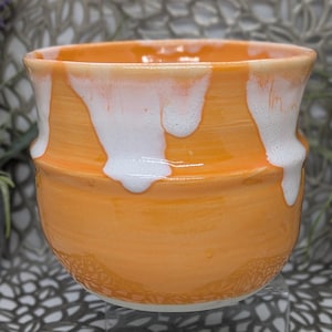 May include: A handmade ceramic bowl with a vibrant orange glaze and white dripped accents. The bowl has a textured base and a slightly flared rim. This unique piece is perfect for serving or as a decorative accent.
