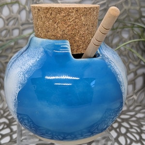 May include: A blue and white ceramic jar with a cork lid and a wooden spoon. The jar has a textured surface and a glossy finish.