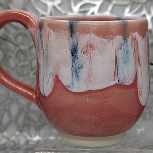 May include: A handmade ceramic mug with a coral-colored exterior and handle. The upper portion features a white glaze with blue and red streaks, creating a unique, dripped effect. The mug sits on a cream-colored base.