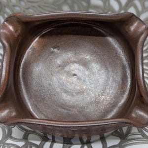 May include: A brown ceramic ashtray with a slightly raised rim and a metallic glaze. The glaze has a swirling pattern and a subtle sheen.