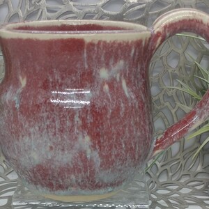 May include: A red ceramic mug with a white glaze and a speckled pattern. The mug has a rounded body and a handle.