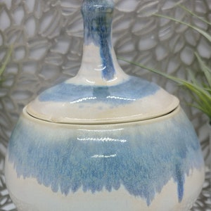 May include: A white ceramic jar with a blue glaze. The jar has a rounded body and a lid with a pointed top. The glaze is uneven, creating a mottled effect.