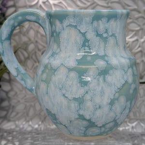 May include: A ceramic mug with a light blue glaze and white, cloud-like patterns. The mug has a curved handle and a slightly flared rim. The base of the mug is a light beige color. The mug is likely handmade.