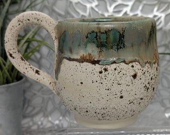 Hand thrown footed 16 oz short porcelain coffee cup in green metallic in places brown and brown speckled cream.