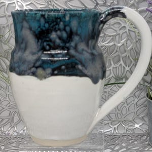 May include: A handmade ceramic mug with a white base and a top section featuring a blend of teal and dark blue glaze. The mug has a large, curved white handle. The mug is designed for hot beverages.