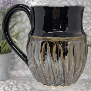 May include: A ceramic mug featuring a glossy black upper section and a textured lower body with vertical fluted grooves. The lower portion displays a metallic, iridescent glaze with bronze and silver tones, separated from the black rim by a band.