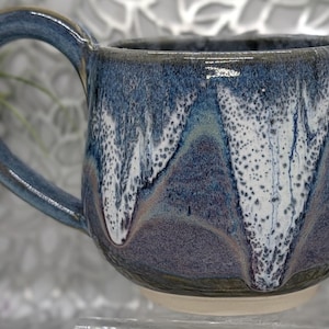 May include: A handmade ceramic mug with a rounded shape and a large handle. The mug features a blue and white glaze with a dripped effect, creating a unique pattern. The base of the mug is a light beige color.