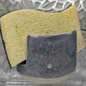 May include: A handmade ceramic sponge holder with a speckled blue and purple glaze, holding a yellow sponge. The holder has a curved design and a light-colored base. The sponge is rectangular with a green border.