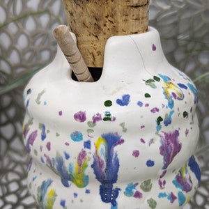 May include: White ceramic jar with a cork lid and a wooden spoon. The jar has a swirl design and is decorated with colorful paint splatters in shades of blue, green, yellow, and purple.