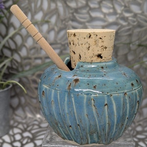 May include: A blue ceramic jar with a cork lid and a wooden spoon. The jar has a textured surface and a rustic look.
