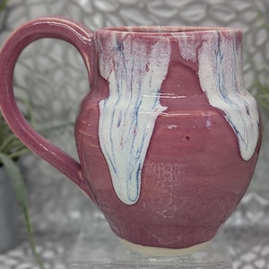 May include: A handmade ceramic mug with a large handle. The mug is a deep rose color with white and blue glaze drips. The mug has a textured surface and a rounded base.
