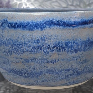 May include: A round ceramic bowl featuring a reactive glaze with horizontal bands of deep cobalt blue and soft grey. The surface displays a textured, dripped effect where the blue glaze blends into the lighter grey base, creating a layered appearance.