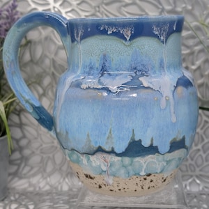 May include: A ceramic mug featuring a rounded body and a curved handle. The glaze displays a layered drip pattern in shades of dark blue, light blue, and white, transitioning to a speckled, unglazed beige base at the bottom.